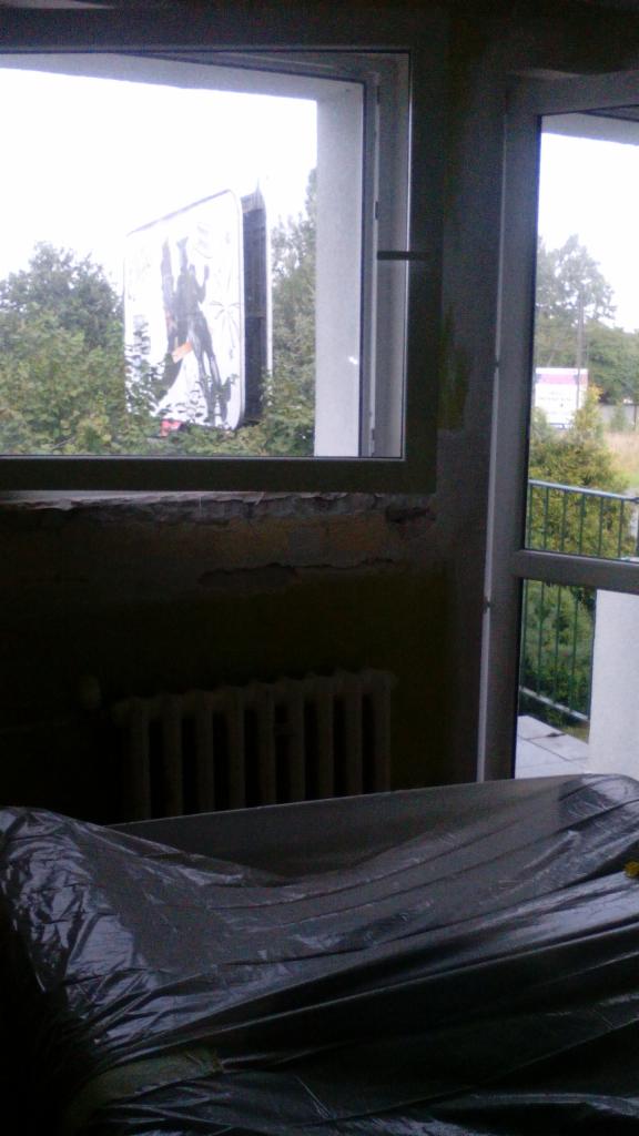 Przygotowania do remontu: Okno z widokiem na zewnątrz, ściana ze śladami starej farby, zakryty folią mebel i grzejnik.