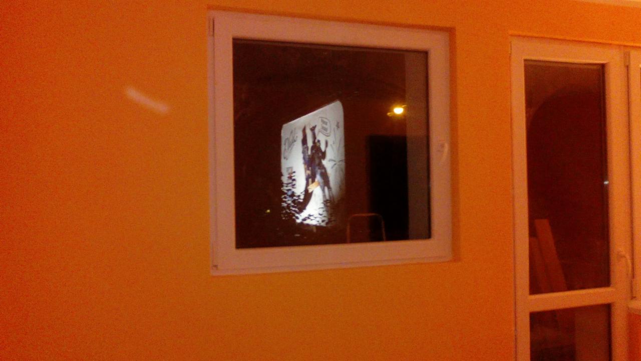 Pomarańczowa ściana z oknem, w którym odbija się rysunek postaci z komiksu na ścianie.