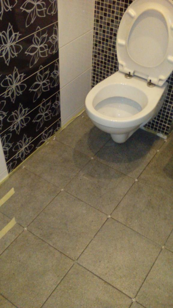 Toaleta z otwartą klapą, częściowo wykończona glazurą na ścianach i szarymi płytkami podłogowymi z krzyżykami dystansowymi, widoczne taśmy malarskie przy ścianie.