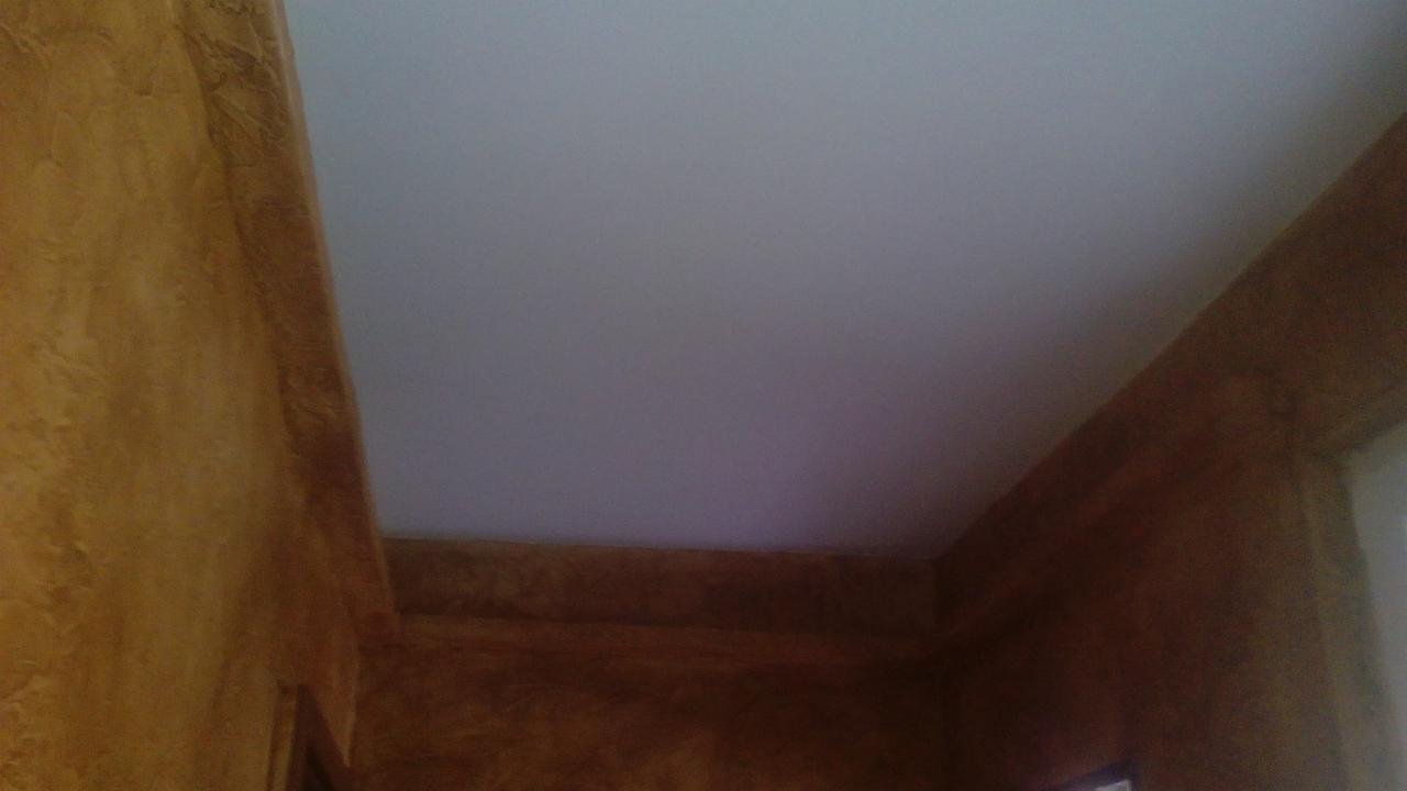 Pomarańczowo-brązowa ściana z wyraźną fakturą i gładki, biały sufit w rogu pokoju, widoczne elementy wykończeniowe.