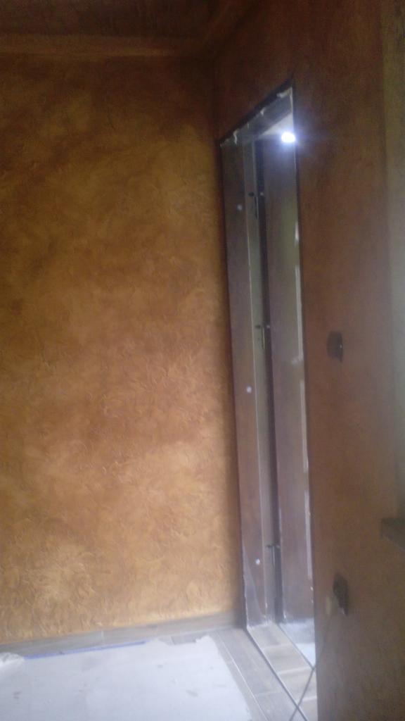 Ściana z dekoracyjną, brązowo-złotą fakturą obok metalowej ościeżnicy drzwiowej bez skrzydła, widoczne fragmenty podłogi wyłożonej płytkami.