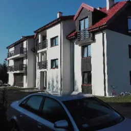 Nowoczesny budynek wielorodzinny z balkonami i czerwoną dachówką, świeżo po remoncie elewacji, z zaparkowanym samochodem na pierwszym planie.