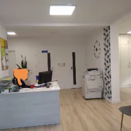 Jasne biuro z recepcją, biurkiem z szufladami, komputerem, drukarką, zegarem ściennym i podłogą z paneli w odcieniach beżu.