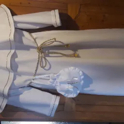 Biała alba komunijna z kapturem i złotym sznurkiem, wykończona złotą lamówką, leżąca na drewnianej szafce.