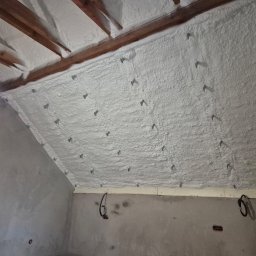 Romel-docieplenia Marcin Kotulak - Poddasze z drewnianą konstrukcją pokryte grubą warstwą białej pianki PUR, zabezpieczone kotwami. Widoczna surowa ściana i oświetlenie robocze.