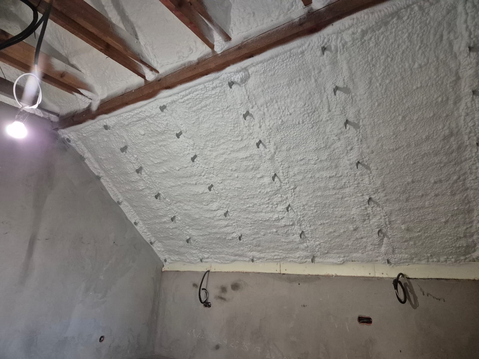 Poddasze z drewnianą konstrukcją pokryte grubą warstwą białej pianki PUR, zabezpieczone kotwami. Widoczna surowa ściana i oświetlenie robocze.
