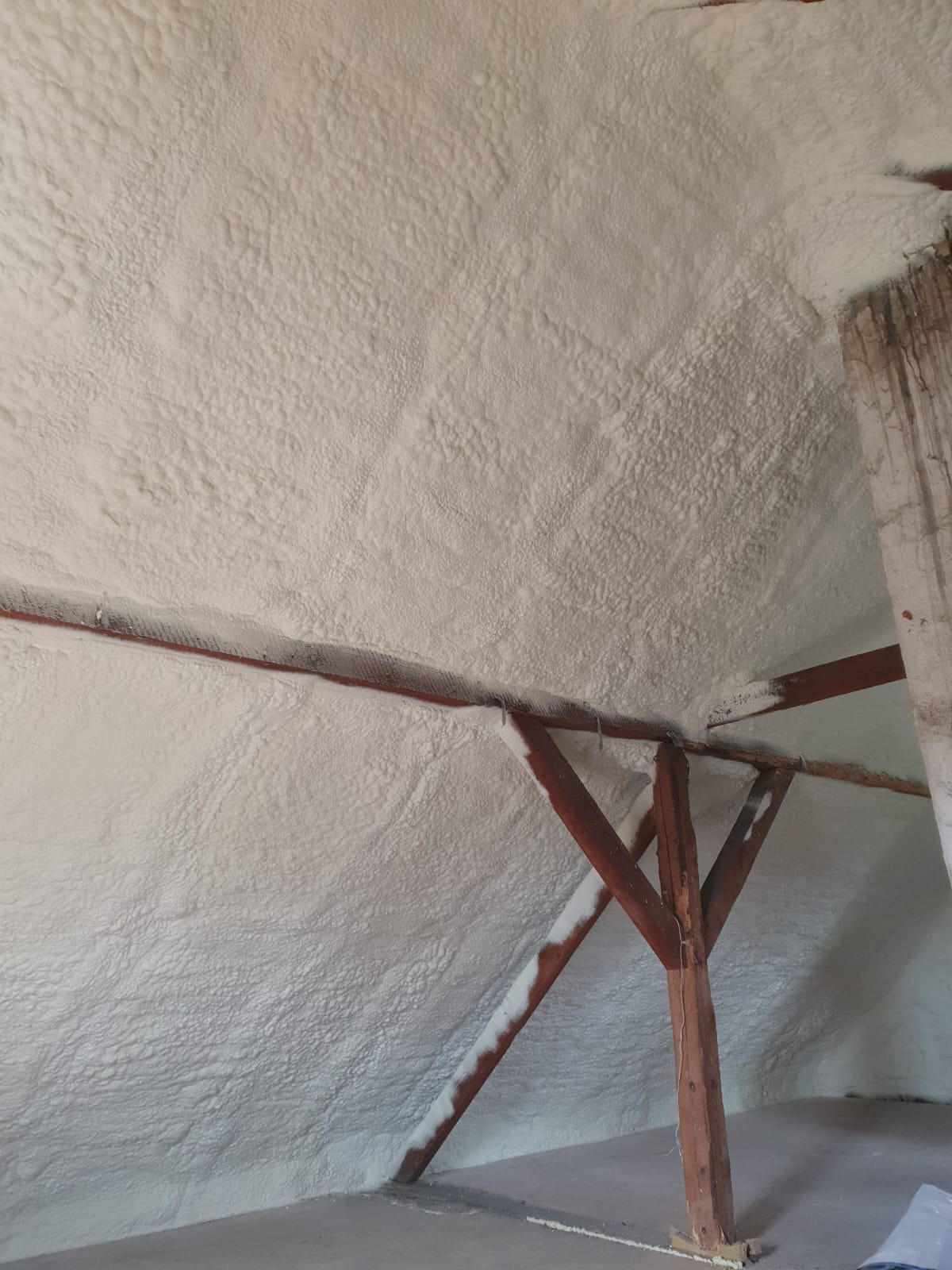 Poddasze po ociepleniu pianką PUR, widoczne drewniane elementy konstrukcyjne. Pianka o nierównej strukturze pokrywa skośne ściany i sufit. Detal wykończenia izolacji.