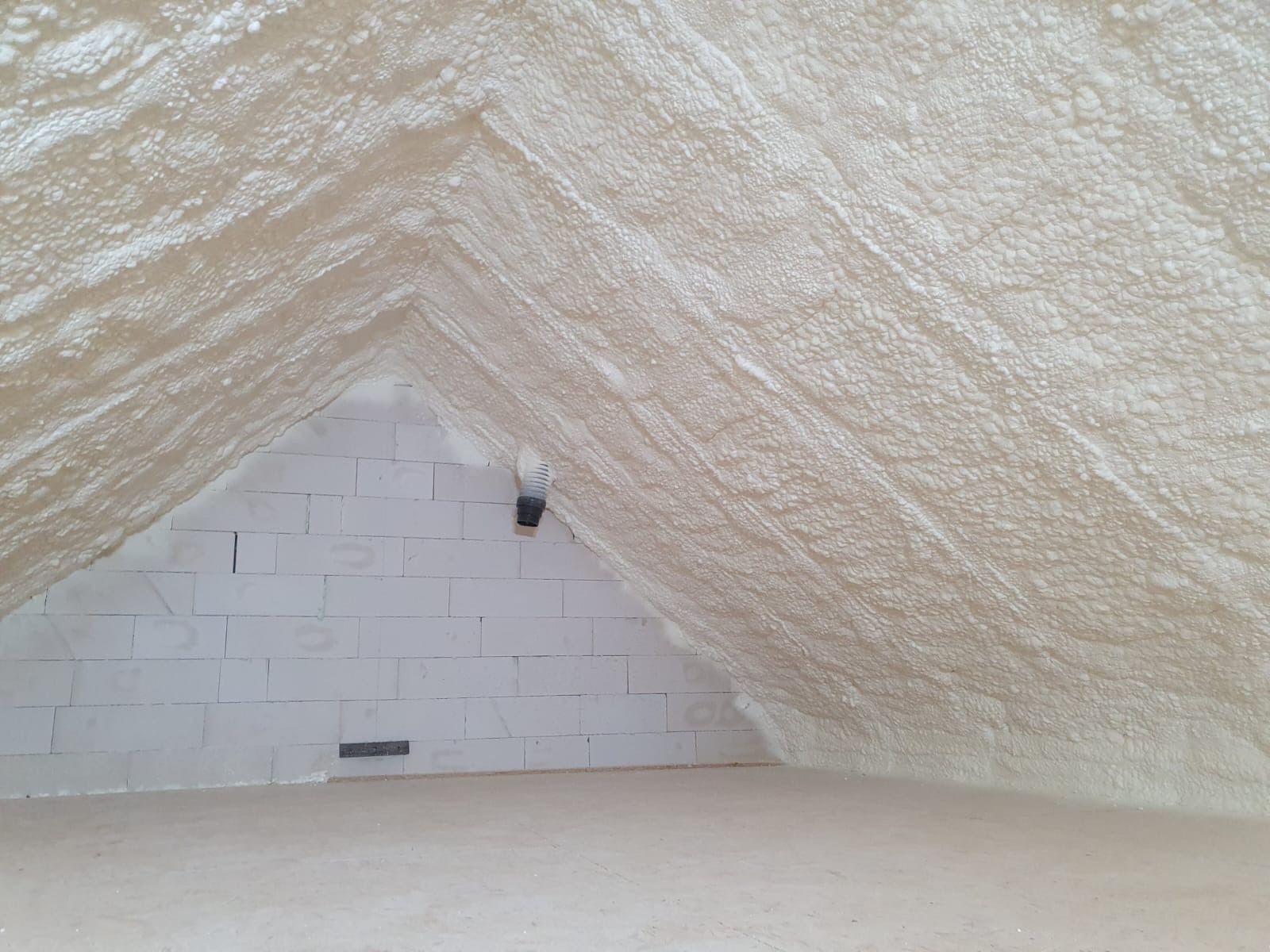 Poddasze po ociepleniu pianką PUR, widoczne białe bloczki i szara rura wentylacyjna. Tekstura pianki przypomina gąbkę, równomiernie pokrywa skosy dachu.