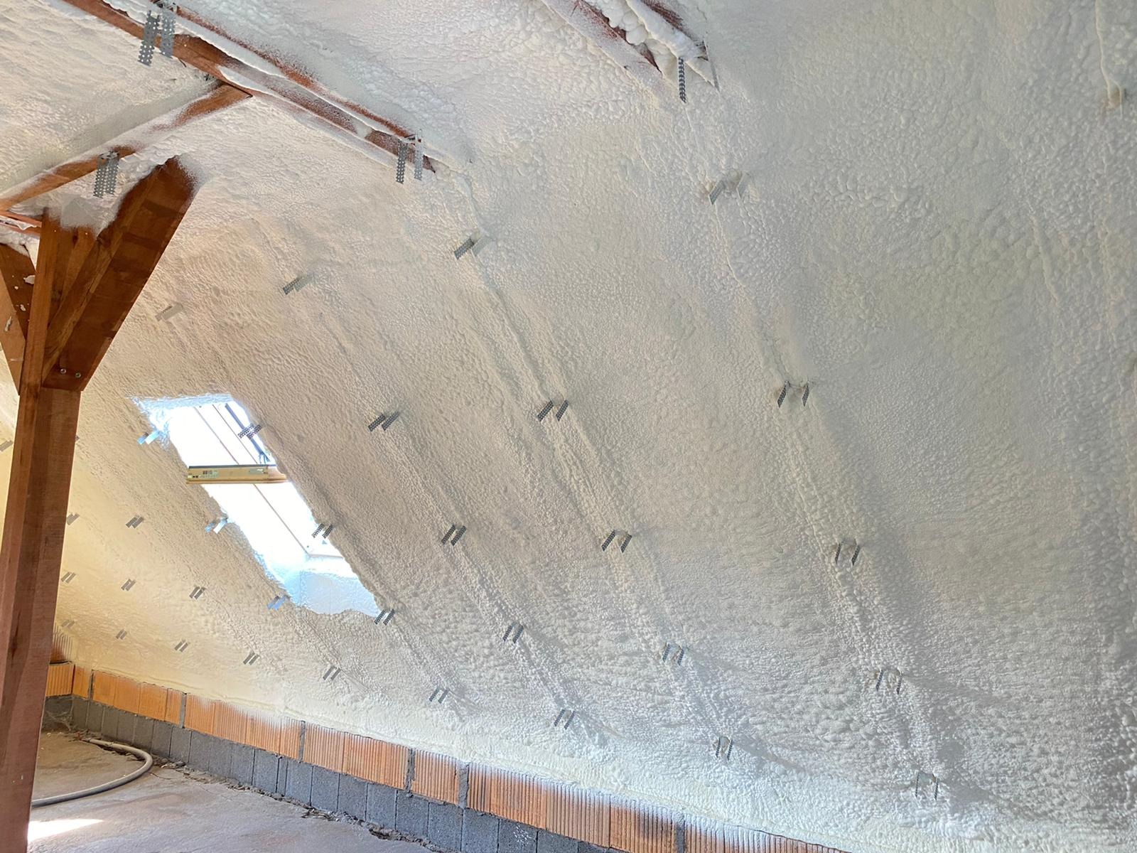 Poddasze z oknem połaciowym po ociepleniu pianką PUR, widoczne drewniane elementy konstrukcyjne i metalowe mocowania izolacji. Biała, nierówna powierzchnia pianki.