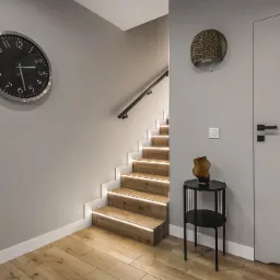 Nowoczesne schody z drewnianymi stopniami podświetlonymi listwami LED, minimalistyczny zegar na szarej ścianie, dekoracyjny koszyk i stolik z wazonem w przedpokoju.