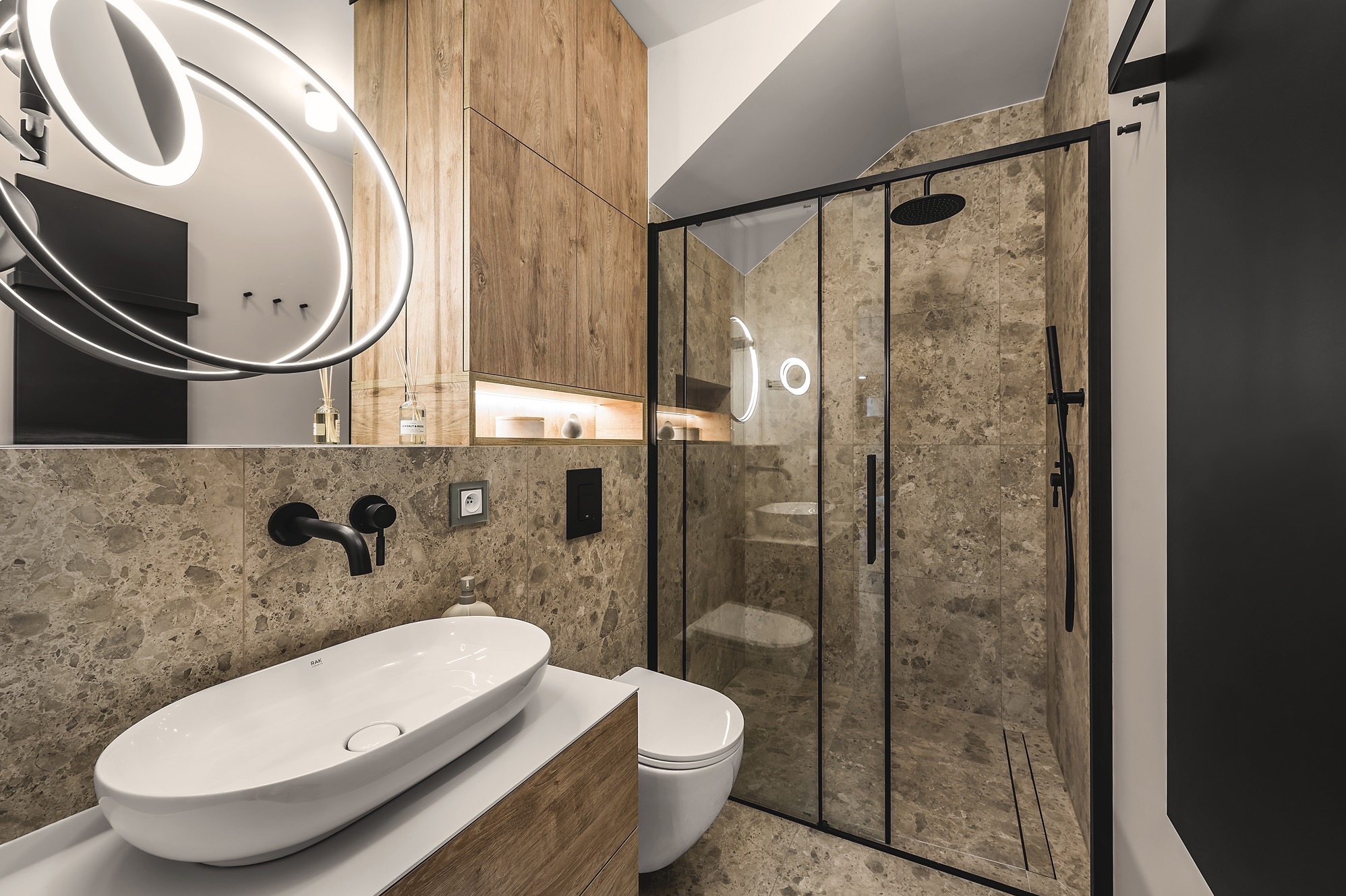 Nowoczesna łazienka z kabiną prysznicową typu walk-in, umywalką nablatową RAK Ceramics, armaturą w kolorze czarnym mat oraz okrągłym lustrem z podświetleniem LED.