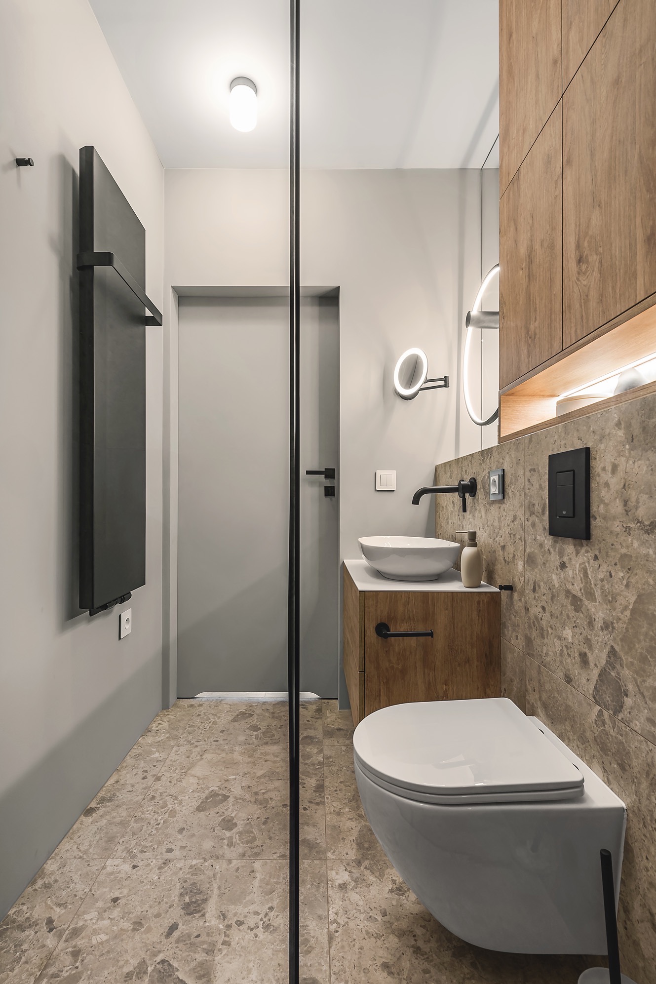 Nowoczesna łazienka z beżowymi płytkami podłogowymi i ściennymi, drewnianą szafką pod umywalką, podwieszaną toaletą i czarnym grzejnikiem drabinkowym.