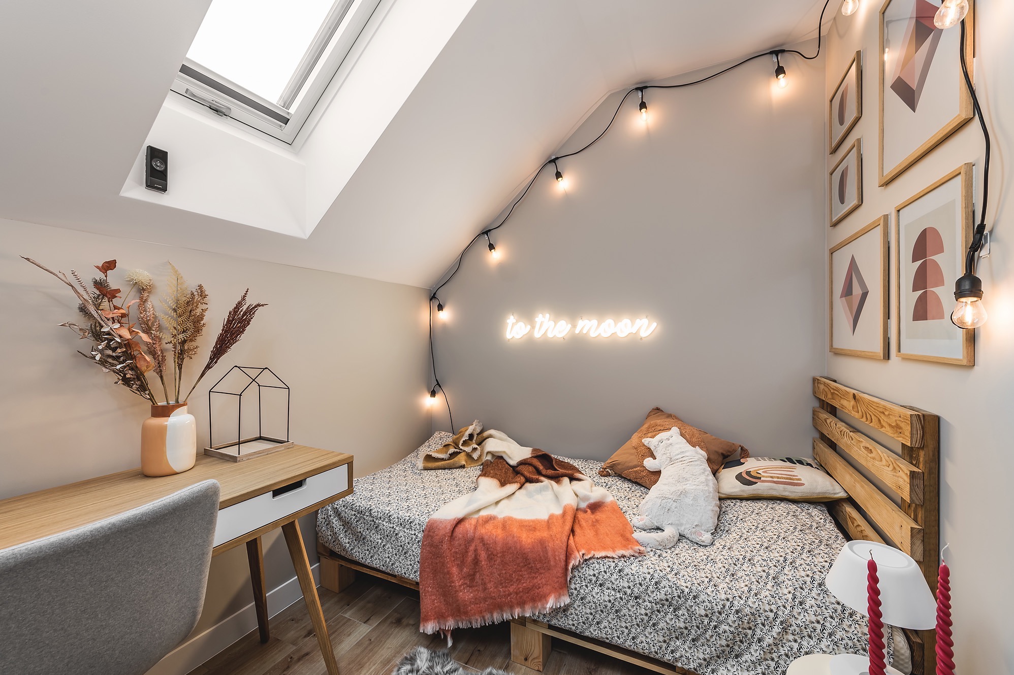 Pokój na poddaszu z łóżkiem na paletach, biurkiem z dekoracjami, oświetleniem girlandą i neonem 'to the moon', oraz oknem dachowym.