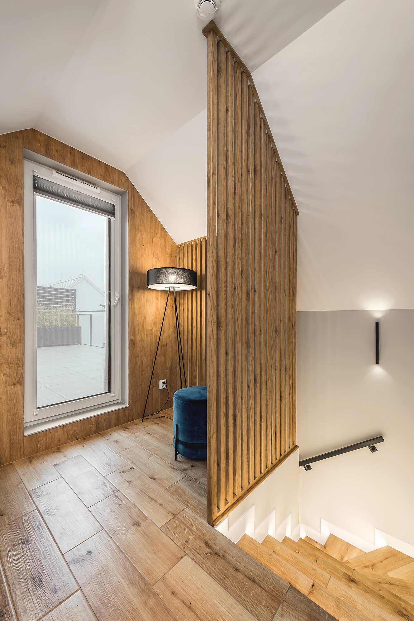 Nowoczesne wnętrze domu z drewnianą ścianą ażurową, podłogą imitującą drewno, schodami z podświetleniem LED, oknem z roletą i turkusowym pufem obok czarnej lampy trójnogu.