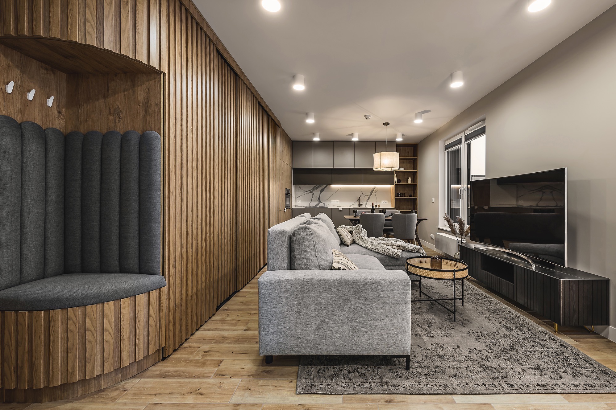 Nowoczesne wnętrze: drewniana ściana z wnęką do siedzenia obitą szarą tkaniną, szara sofa z poduszkami, dywan w odcieniach szarości, telewizor na szafce RTV i widoczna w tle kuchnia z białym blatem...