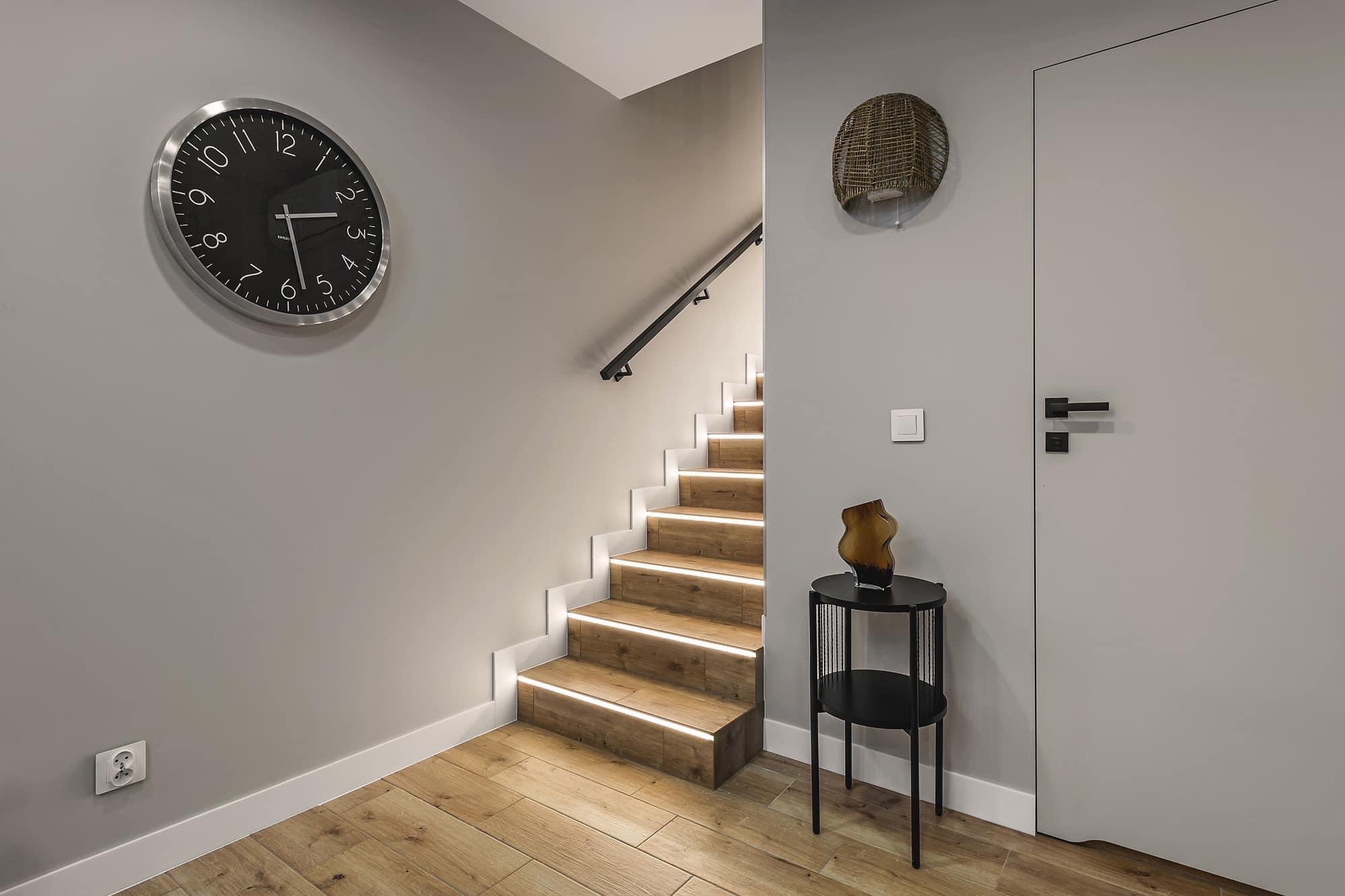 Nowoczesne schody z drewnianymi stopniami podświetlonymi listwami LED, minimalistyczny zegar na szarej ścianie, dekoracyjny koszyk i stolik z wazonem w przedpokoju.