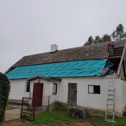 Piotr Kośmiński - Dom w trakcie wymiany dachu. Widoczny dekarz na połowie dachu, zabezpieczonej folią FAKRO, oraz sterta dachówek obok ściany. Drabina oparta o budynek.