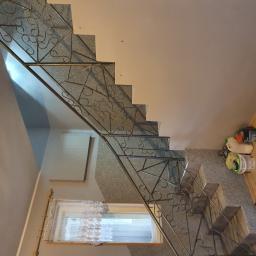 Piotr Kośmiński - Granitowe schody z metalową balustradą o dekoracyjnym wzorze, widok z góry. Obok okna firanka z koronkowym wzorem. Ściany pomalowane na jasny kolor.