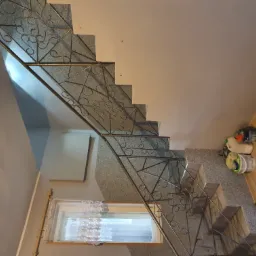 Granitowe schody z metalową balustradą o dekoracyjnym wzorze, widok z g&oacute;ry. Obok okna firanka z koronkowym wzorem. Ściany pomalowane na jasny kolor.