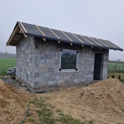 Piotr Kośmiński - Budynek gospodarczy w trakcie budowy: ściany z szarego bloczka, wstępne krycie dachu papą, okno z PCV, hałda piasku na pierwszym planie.