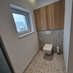 Piotr Kośmiński - Nowoczesna toaleta z drewnianą zabudową nad stelażem, mozaikową ścianą i podłogą w stylu lastryko, oświetlona światłem z okna i lampy sufitowej.