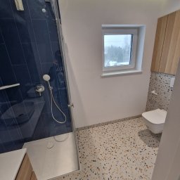 Piotr Kośmiński - Nowoczesna łazienka z kabiną prysznicową typu walk-in, granatowymi płytkami na ścianie, umywalką z drewnianą szafką, toaletą wiszącą i podłogą w stylu lastryko.