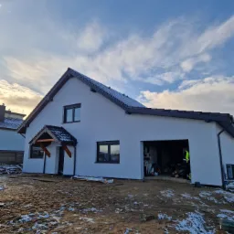 Nowoczesny dom jednorodzinny z ciemną dachówką i jasną elewacją, widok z zewnątrz, teren wokół domu częściowo pokryty śniegiem.