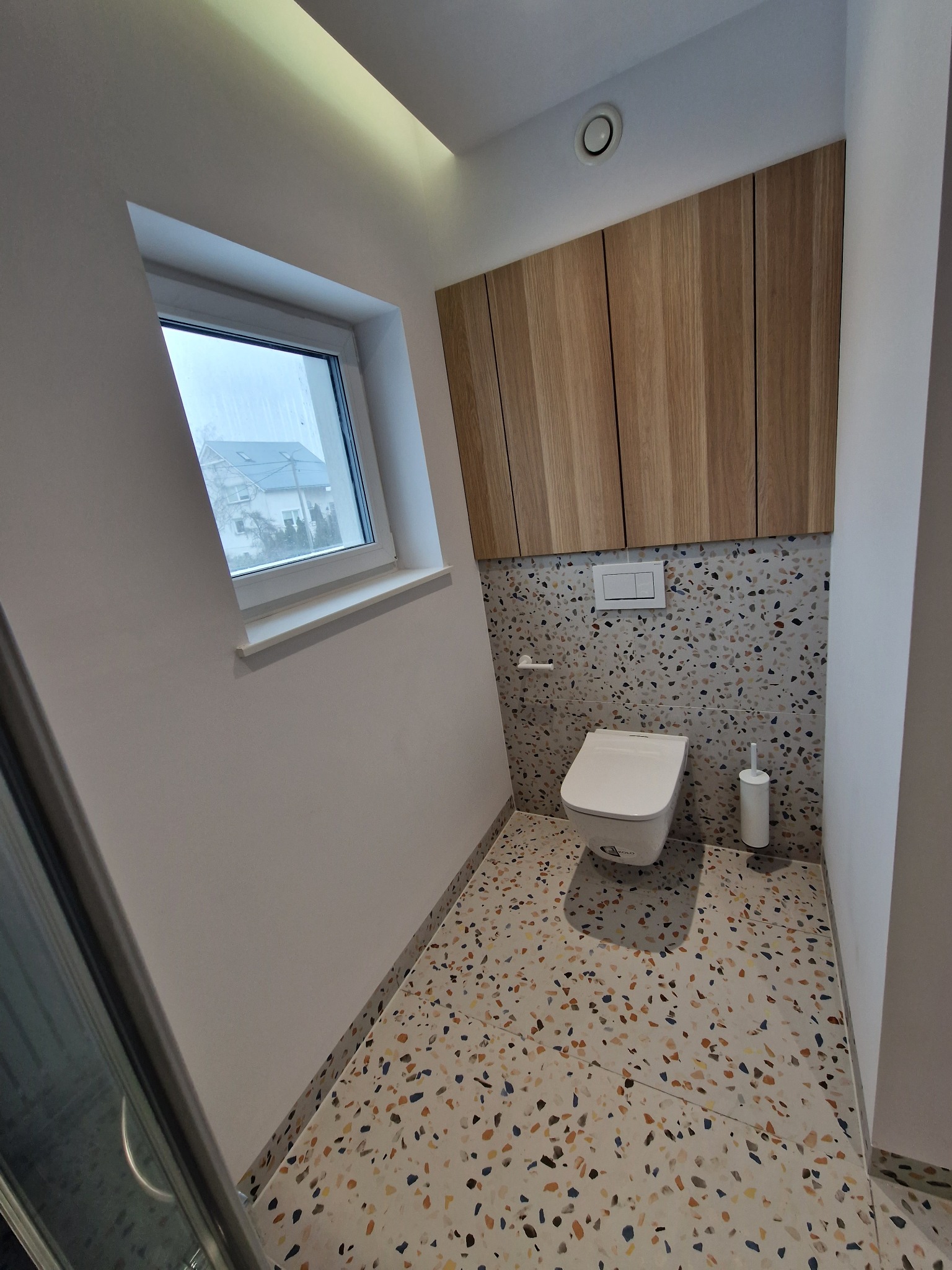 Nowoczesna toaleta z drewnianą zabudową nad stelażem, mozaikową ścianą i podłogą w stylu lastryko, oświetlona światłem z okna i lampy sufitowej.