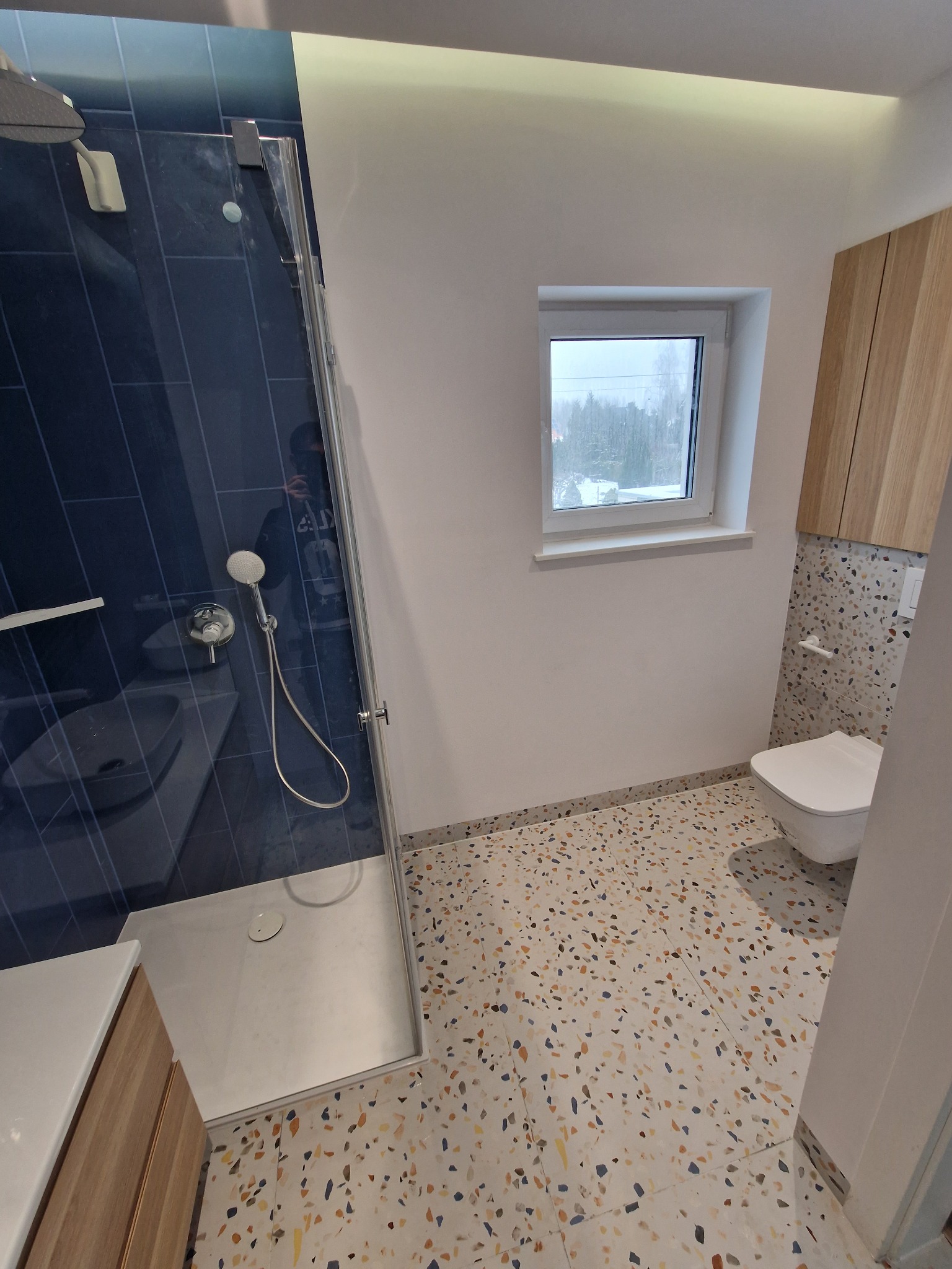 Nowoczesna łazienka z kabiną prysznicową typu walk-in, granatowymi płytkami na ścianie, umywalką z drewnianą szafką, toaletą wiszącą i podłogą w stylu lastryko.