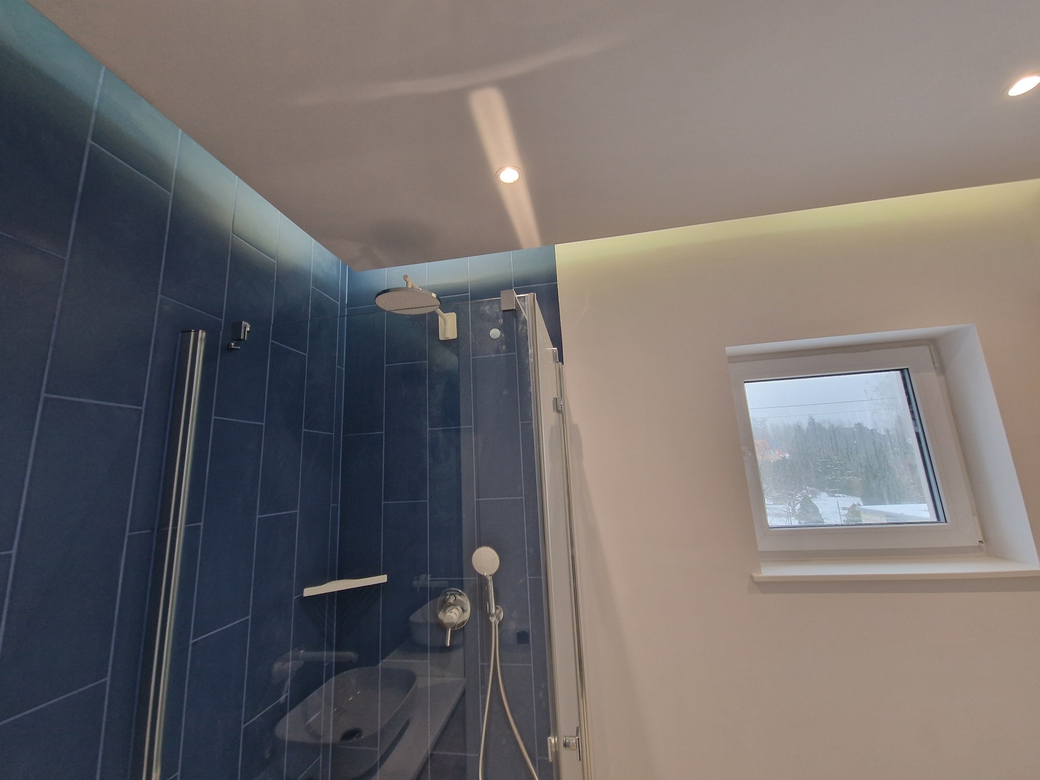 Nowoczesna kabina prysznicowa z deszczownicą i baterią natryskową na tle ściany wyłożonej ciemnoniebieskimi, pionowymi płytkami; widok na zewnątrz przez okno dachowe.