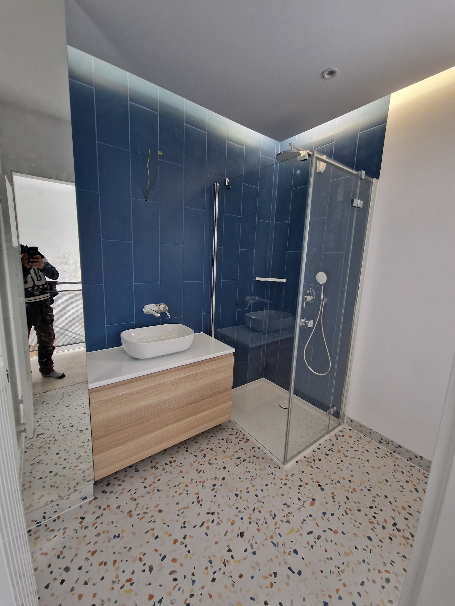 Nowoczesna łazienka z kabiną prysznicową, umywalką nablatową na szafce z drewnopodobnym fornirem, niebieskimi płytkami na ścianie i podłogą w stylu lastryko. Widoczne wystające przewody elektryczne.