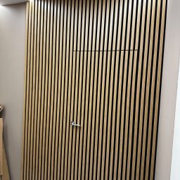 Stet-Tech - Ukryte drzwi wykończone pionowymi listwami drewnianymi, z minimalistyczną klamką i ukrytym panelem dostępowym, wkomponowane w ścianę z oświetleniem LED przy suficie.
