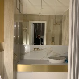 Stet-Tech - Nowoczesna łazienka z umywalką nablatową, złotymi akcentami na szafkach i armaturze, oraz lustrami panelowymi na ścianie, w których odbija się osoba robiąca zdjęcie.
