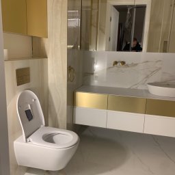 Stet-Tech - Nowoczesna, podwieszana toaleta z otwartą klapą w łazience z marmurowymi ścianami i złotymi akcentami, widoczne złote szafki i umywalka nablatowa, odbicie osoby w lustrze.