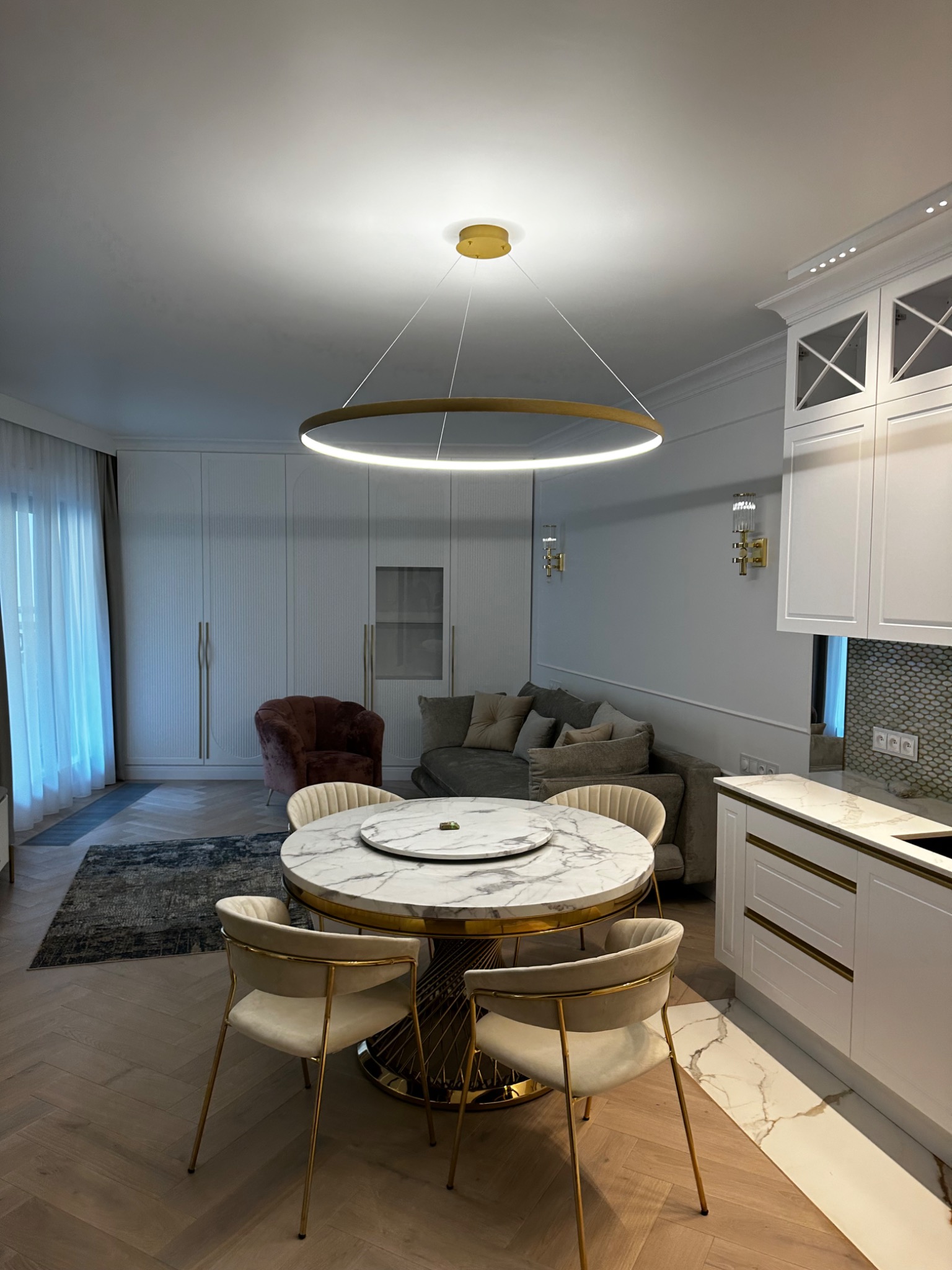Nowoczesne wnętrze z okrągłym stołem z marmurowym blatem i złotą podstawą, krzesła tapicerowane beżowym welurem z metalowymi złotymi nogami, wisząca okrągła lampa LED, biała zabudowa kuchenna...