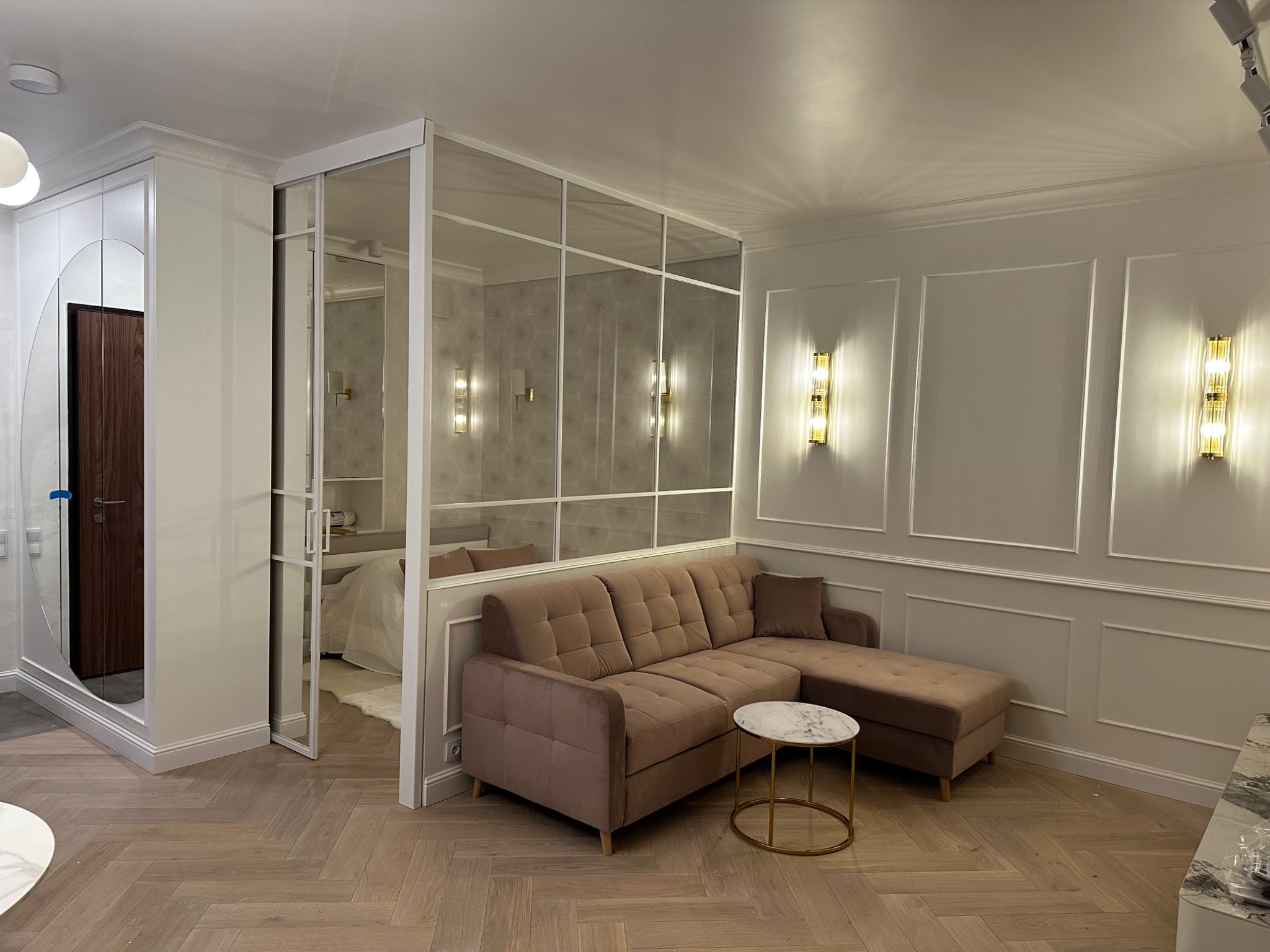 Elegancki salon z beżową sofą i stolikiem kawowym na parkiecie ułożonym w jodełkę, oddzielony szklaną ścianą od sypialni, z dekoracyjnymi lampami ściennymi.