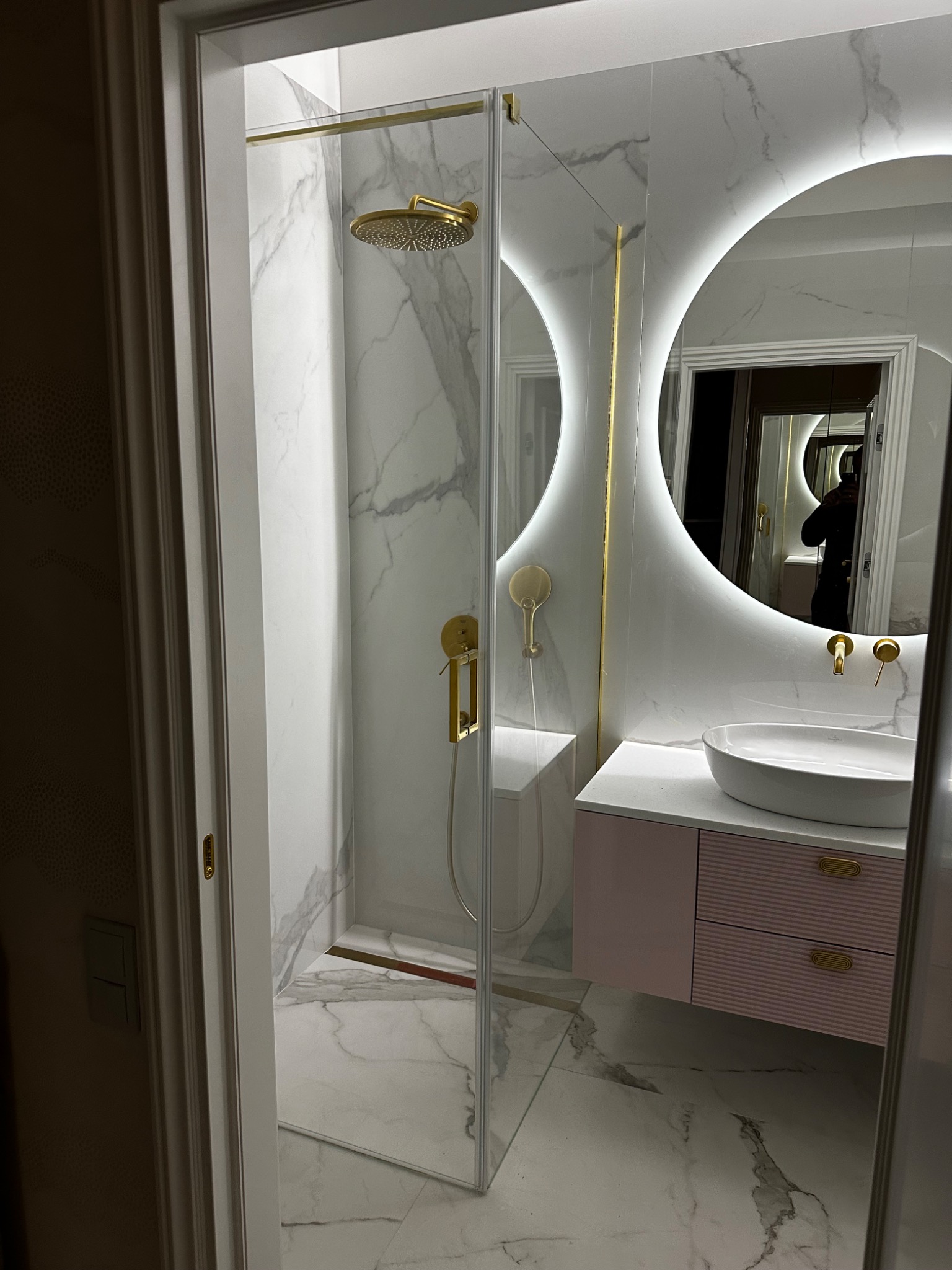 Nowoczesna łazienka z kabiną prysznicową typu walk-in, marmurowymi płytkami, okrągłym lustrem z podświetleniem LED i umywalką nablatową na szafce w odcieniach różu.