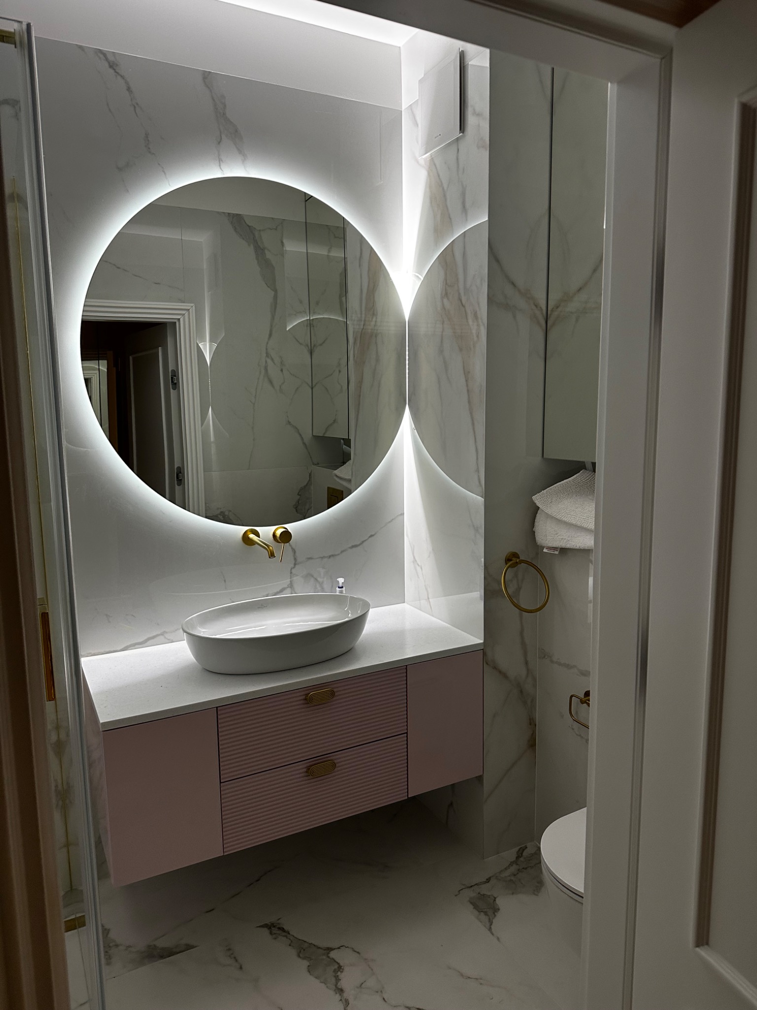 Nowoczesna łazienka z okrągłym lustrem podświetlanym LED, białą umywalką nablatową na różowej szafce z poziomymi frezami, złotą armaturą i marmurowymi płytkami na ścianach i podłodze.