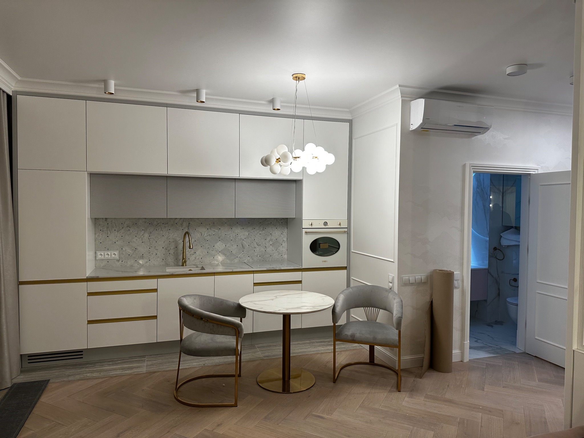 Nowoczesna, jasna kuchnia z białymi szafkami z delikatnymi złotymi akcentami, marmurowym blatem i okrągłym stołem z dwoma szarymi krzesłami na parkiecie ułożonym w jodełkę; widok na łazienkę w tle...