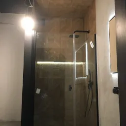 Nowoczesna kabina prysznicowa z czarną armaturą i szklaną ścianką, drewnopodobne płytki na ścianach i suficie ułożone we wzór jodełki, oświetlenie LED w lustrze i pod prysznicem, widoczne...