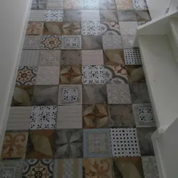 Wzór podłogi z kwadratowych płytek ceramicznych w odcieniach beżu, szarości i brązu, ułożonych w patchworkowy deseń, widoczne z perspektywy korytarza przy schodach.