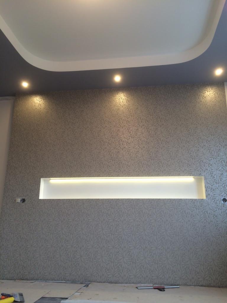 Ściana z szarą tapetą w kwiaty, oświetlona wnęka z listwą LED, sufit podwieszany z oświetleniem punktowym, widoczne narzędzia na podłodze.
