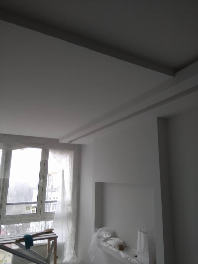 Białe wnętrze w trakcie prac wykończeniowych: widoczne sufity podwieszane z dekoracyjnymi listwami, okno z osłoną malarską i prowizoryczny stolik roboczy z narzędziami.