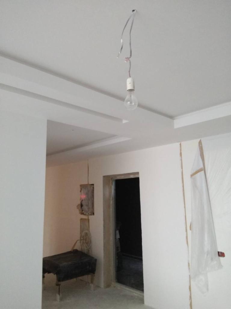 Pokój w trakcie odświeżania: świeżo pomalowane ściany na biało, widoczne sztukaterie sufitowe, wisząca żarówka na kablu, niezabezpieczona skrzynka elektryczna i zakryte folią drzwi.