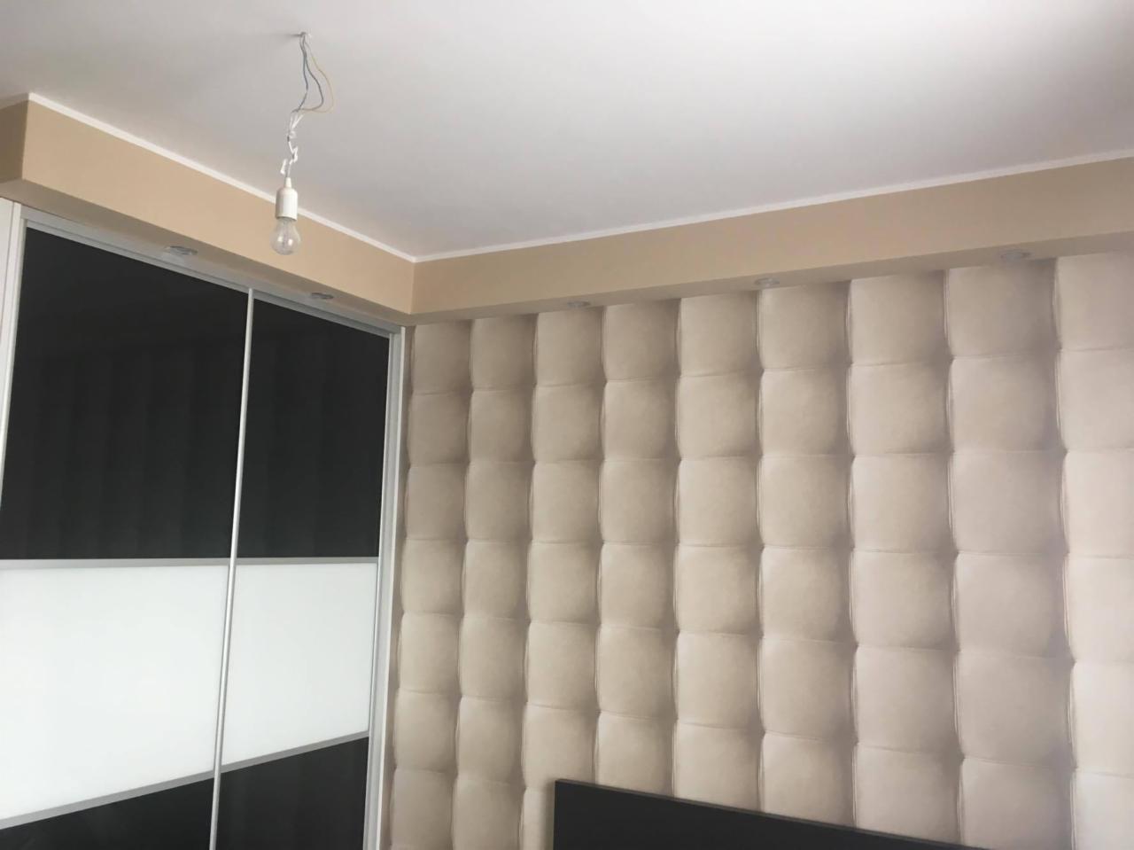 Tapicerowany panel ścienny w beżowym odcieniu, widoczny fragment sufitu z niedokończonym oświetleniem i przesuwna szafa z czarno-białymi drzwiami.