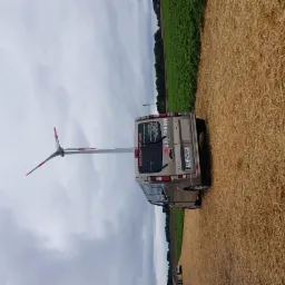 Beżowy bus firmy transportowej z numerem telefonu i nazwą 'KRZYSZTOF-BUS' zaparkowany na polu po żniwach, z turbiną wiatrową w tle.