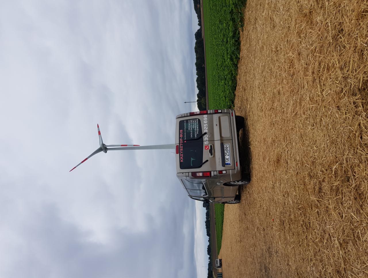 Beżowy bus firmy transportowej z numerem telefonu i nazwą 'KRZYSZTOF-BUS' zaparkowany na polu po żniwach, z turbiną wiatrową w tle.
