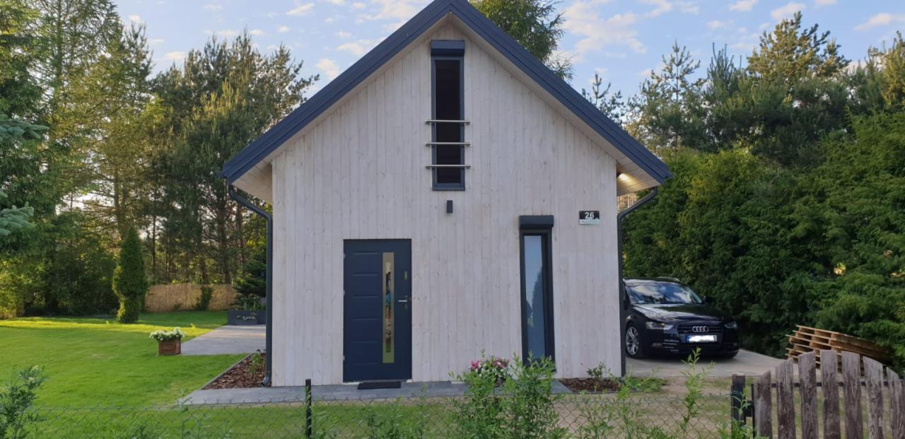 Nowoczesny dom szkieletowy z jasną elewacją, ciemnymi ramami okiennymi i drzwiami, numerem domu '26' oraz samochodem osobowym zaparkowanym obok