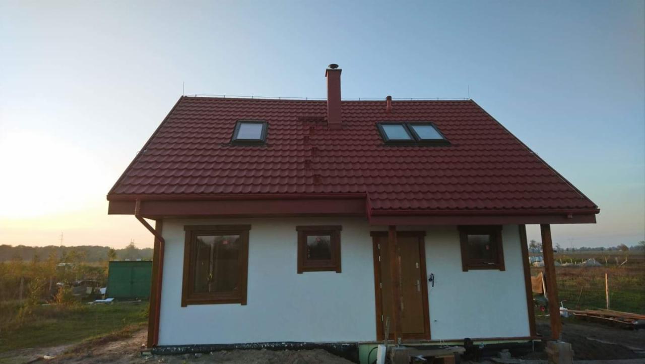 Nieduży dom z czerwonym dachem z dachówki, widoczne okna dachowe, komin i brązowe ramy okien i drzwi, na tle pola i lasu.