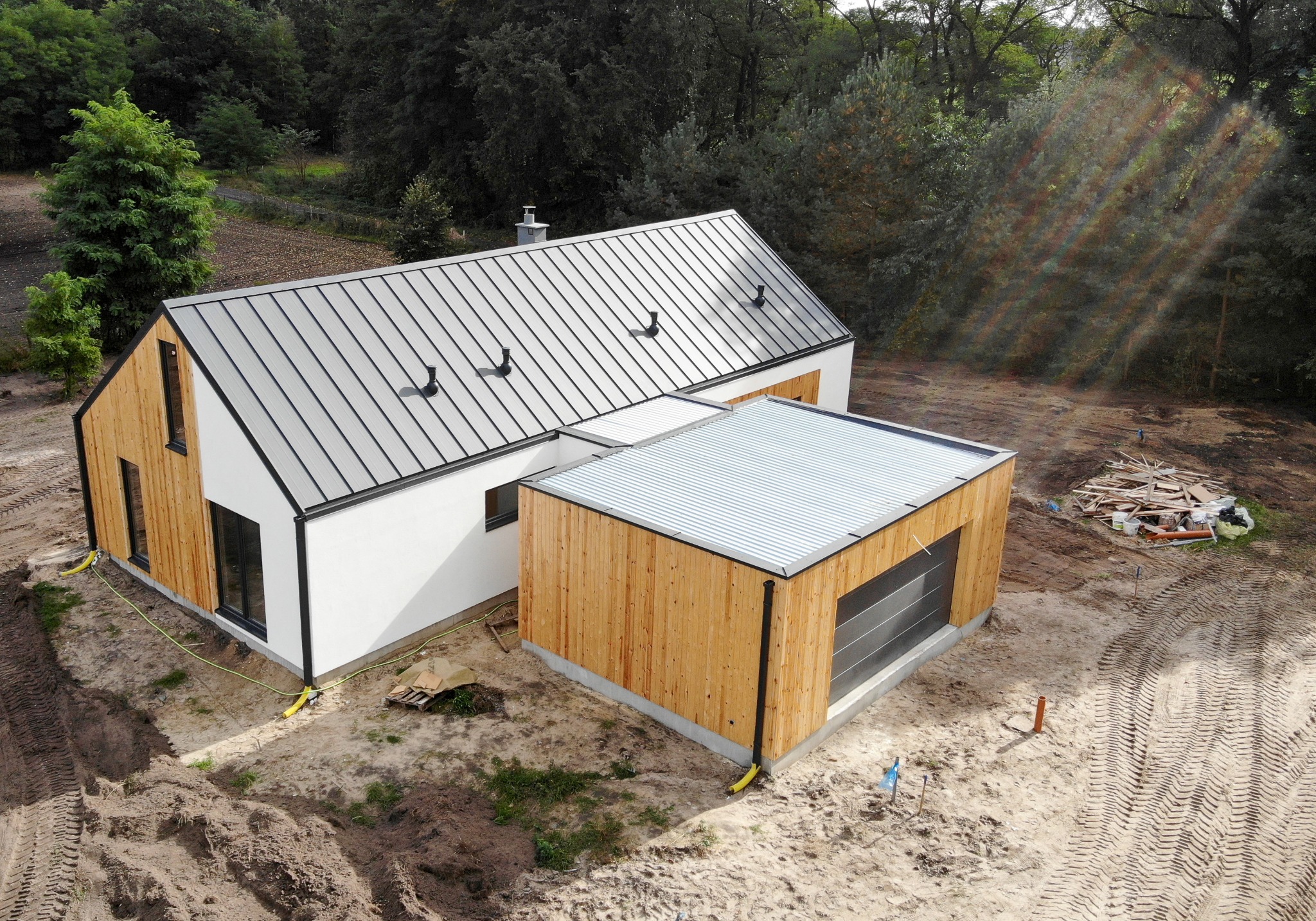 Nowoczesny dom z drewnianą elewacją i metalowym dachem, widok z góry, teren budowy z widocznymi śladami opon i stertą drewna.